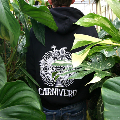 Back view of a person wearing a black hoodie with a white Carnivero logo among large tropical leaves
